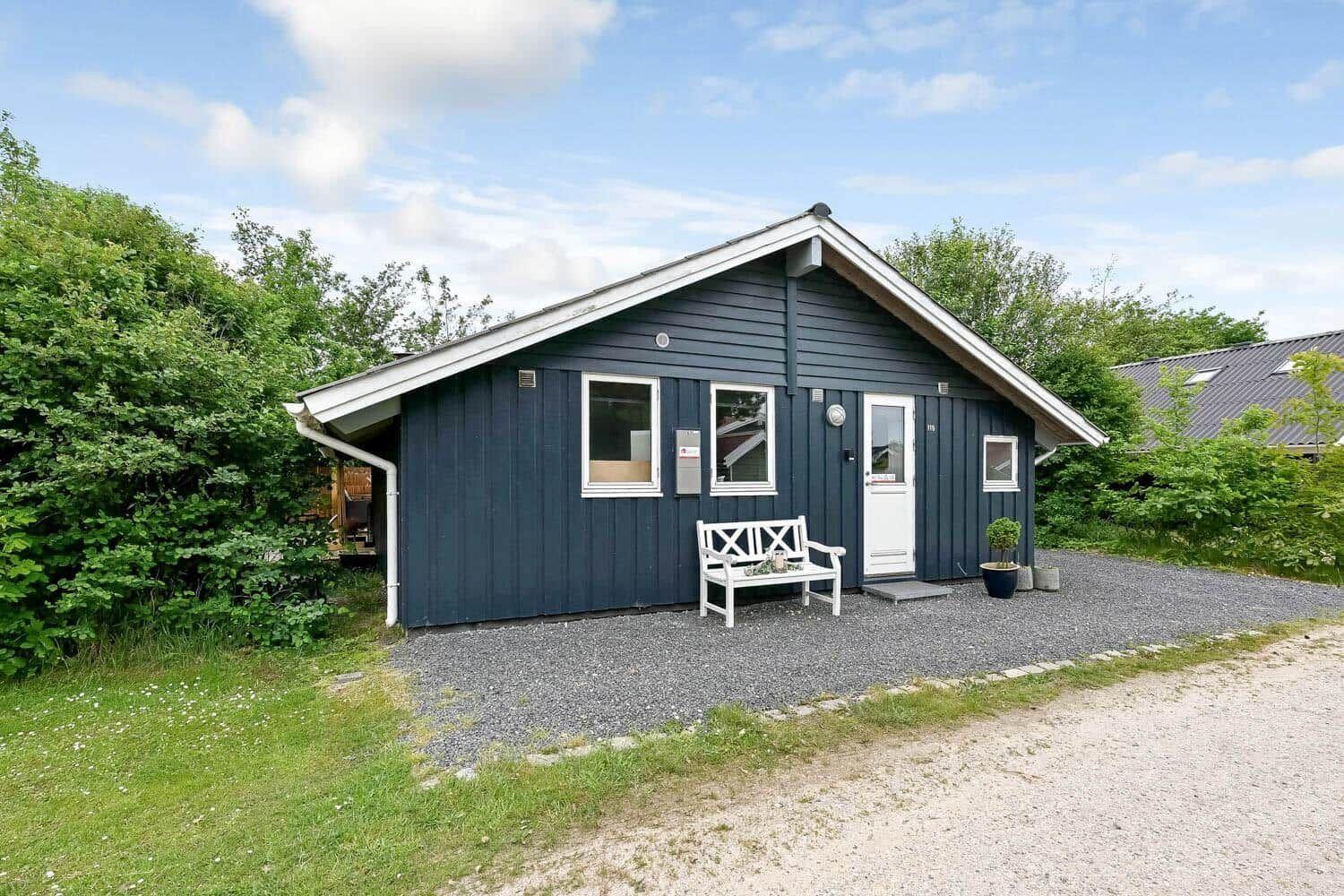 Ein dunkelblaues Holzhaus mit weißer Tür und Bank vor dem Eingang. Gras und Bäume umgeben das Grundstück.