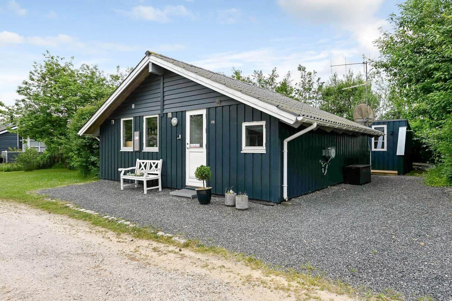 Ein dunkelblaues Ferienhaus mit weißer Tür und Bank vorne. Gras und Bäume umgeben es.