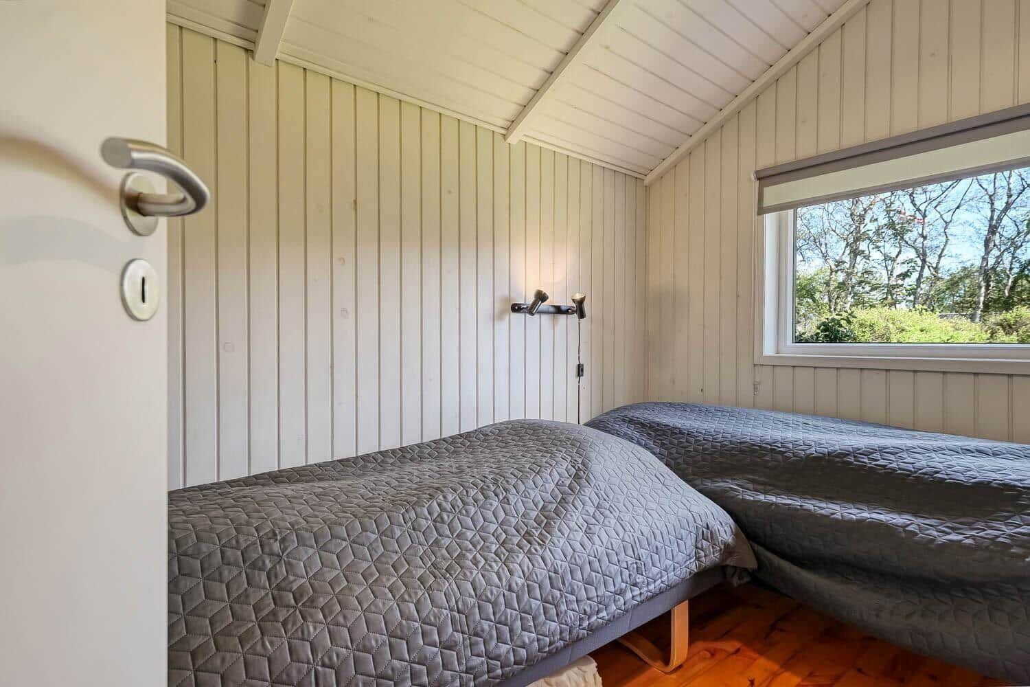 Two beds with gray covers in a room with wood paneling and a window.