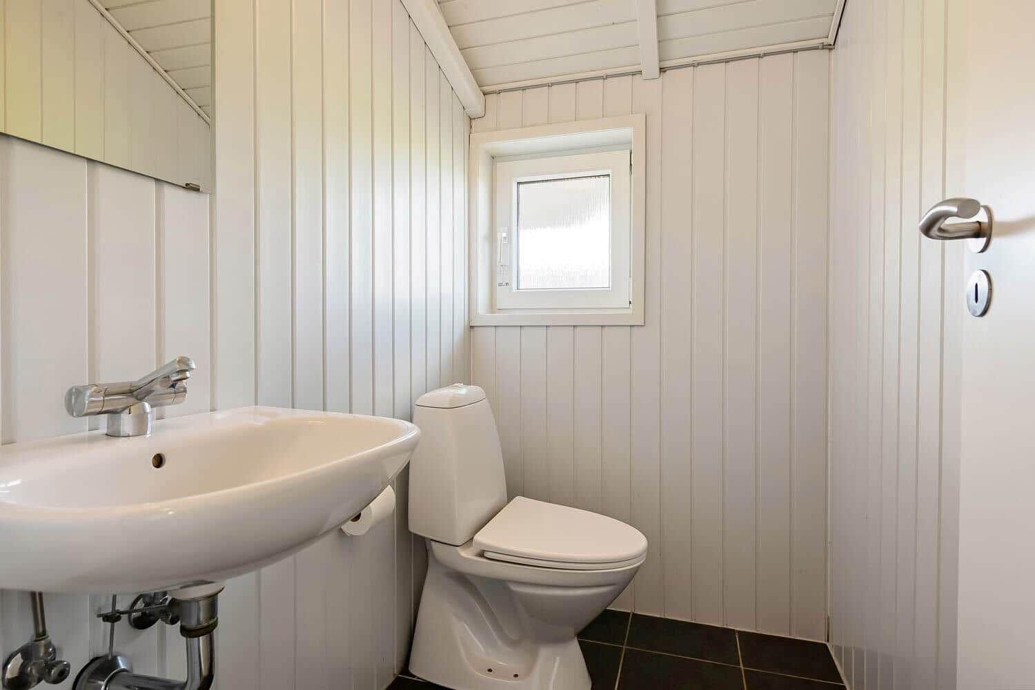 Bathroom with toilet, sink, and window. Walls are clad in white paneling.