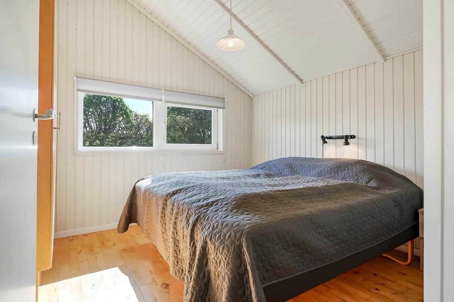 Bedroom with bed, window, and wooden floor. Sloped ceiling, white panel walls.