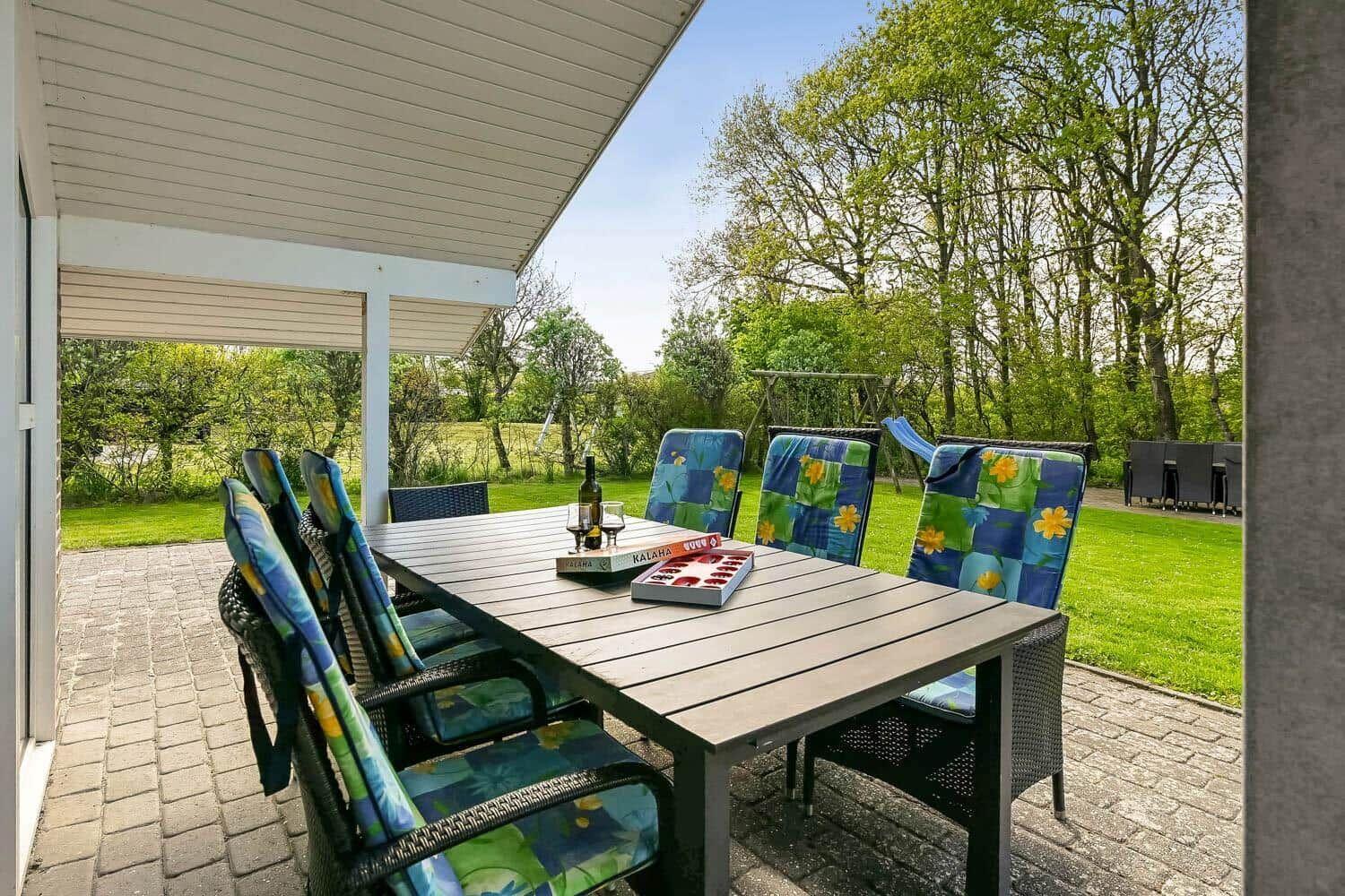 Patio with table and chairs under roof. Background: lawn and trees.