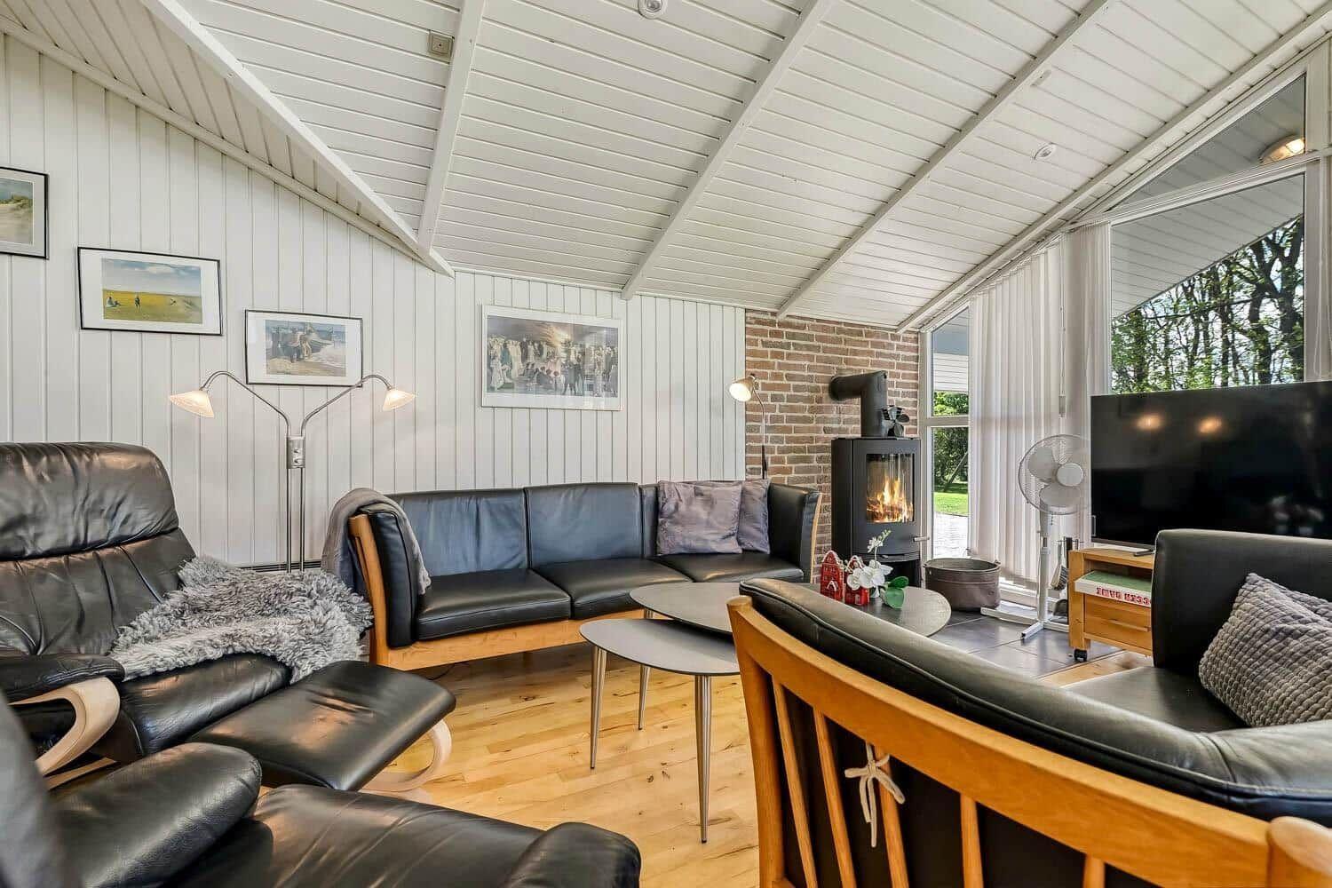 Living room with sofa, armchairs, and wood-burning stove. Wooden floor and large windows.