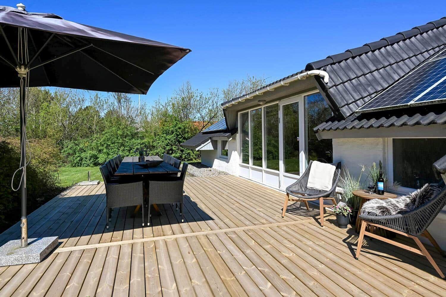Terrasse mit Tisch, Stühlen und Sonnenschutz. Hintergrund: Haus mit Solarpanel und Garten.
