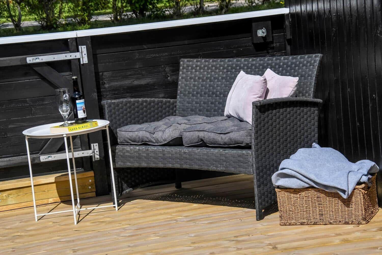 Terrasse mit Gartenbank, Tisch, Flasche und Kissen.