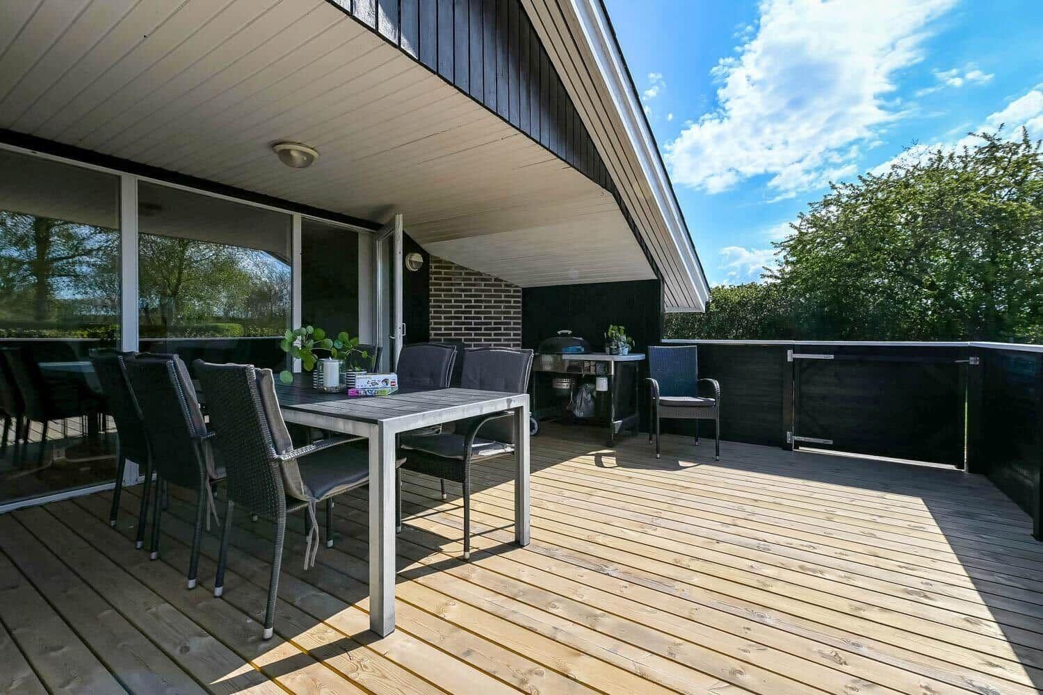 Terrasse mit Tisch, Stühlen und Grill unter Dach. Holzdecke, Sicht auf Bäume.