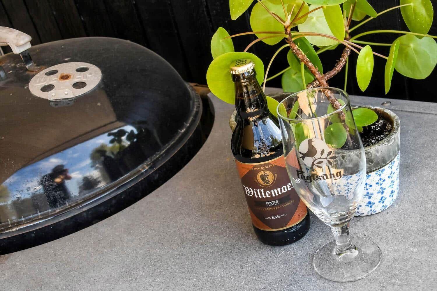 Eine Flasche Bier und ein Glas stehen auf einer Terrasse neben einem Grill und einer Pflanze.