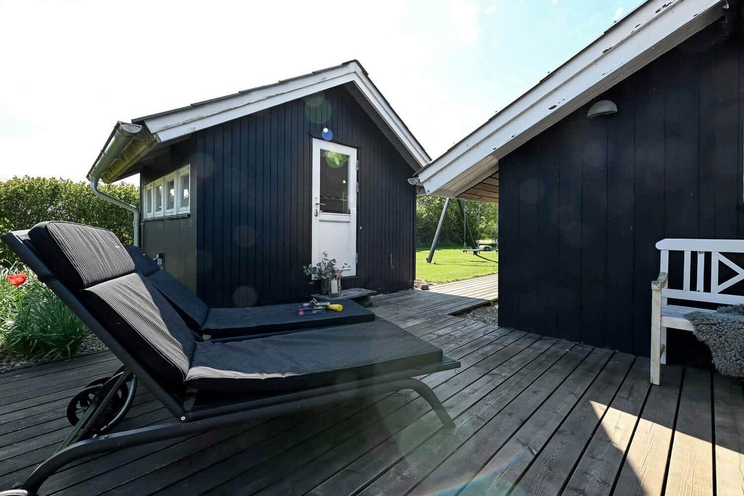 Schwarze Holzhütte mit Terrasse, Liegestuhl und weißem Stuhl.