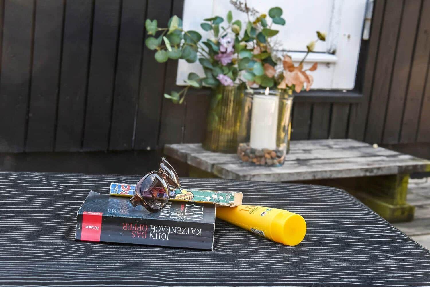 Auf einem Tisch liegen Buch, Sonnenbrille und Sonnenschutzcreme. Im Hintergrund Blumen und Kerze.