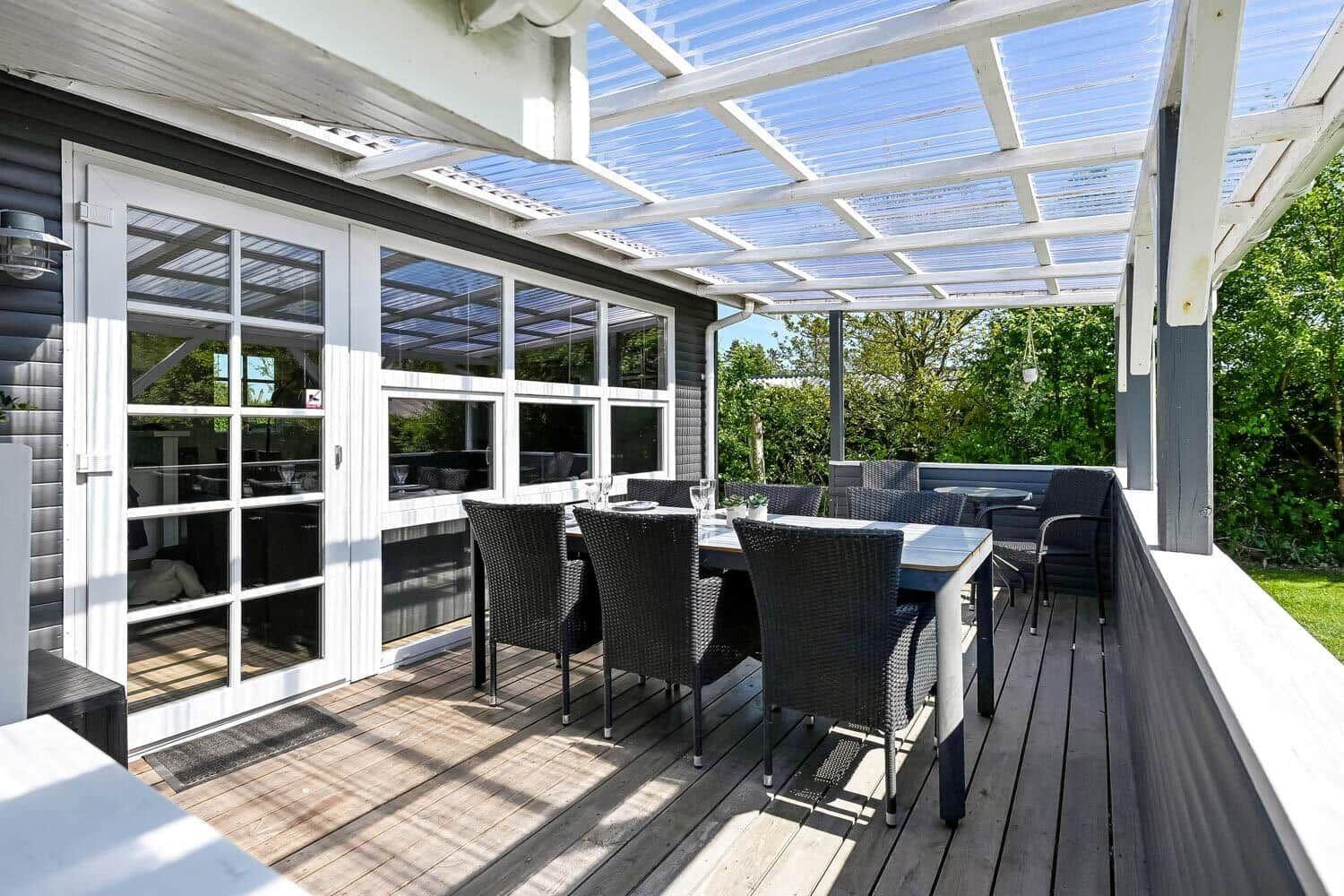 Terrasse mit Tisch, Stühlen und Dachkonstruktion. Durchgehendes Glasfenster zur Inneneinrichtung.