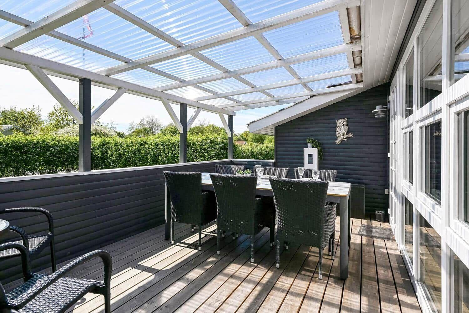 Terrasse mit Tisch und Stühlen unter Glasdach. Holzdecke, dunkle Wand und grüner Hintergrund.