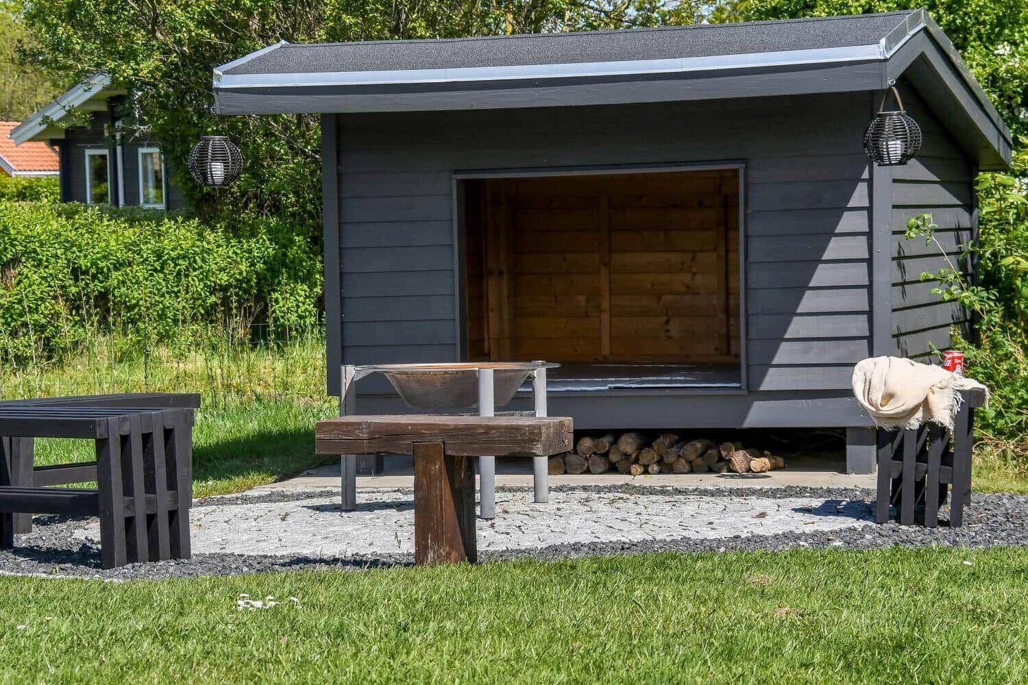 Ein graues Holzhaus mit offener Tür und Brennholz darunter. Draußen steht ein Tisch und Bänke.