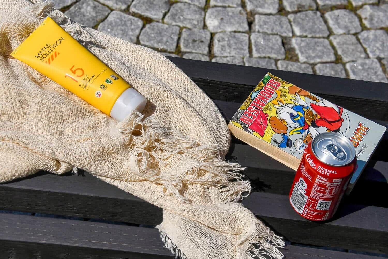 Sonnencreme, Getränk und Comic auf einer Bank vor Steinplatten.