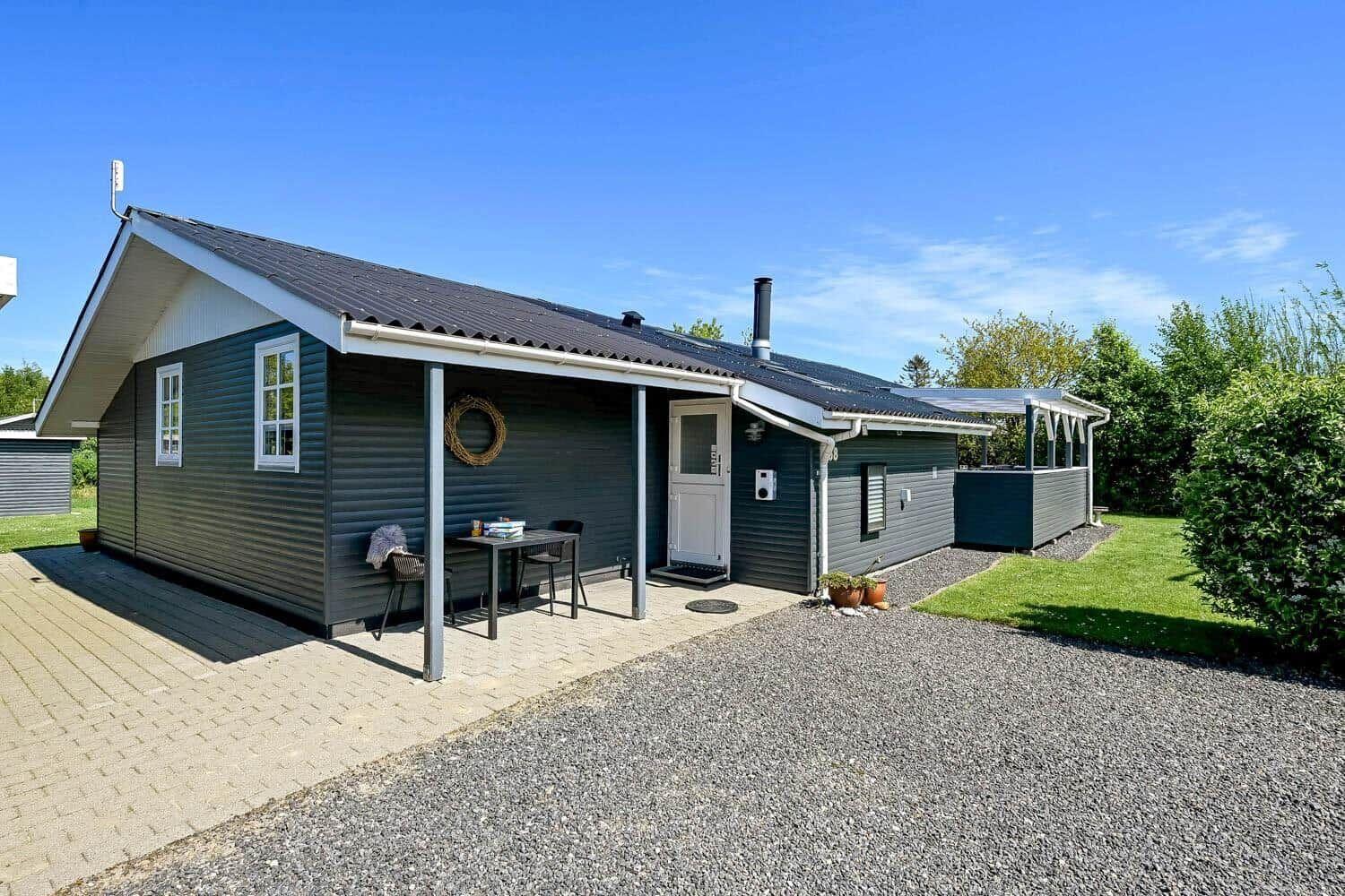 Ein graues Ferienhaus mit Terrasse, Garten und Grillplatz unter blauem Himmel.