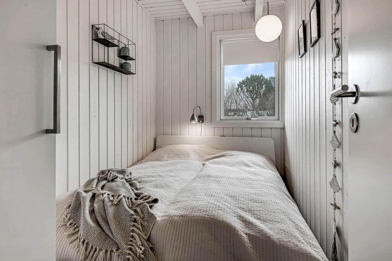 Ein Schlafzimmer mit weißer Holzwand, Bett und Fenster mit Blick auf Bäume.