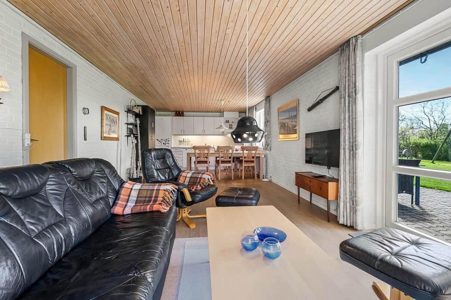 Wohnzimmer mit Ledersofa, Essbereich und TV. Holzdecke und große Fenster.