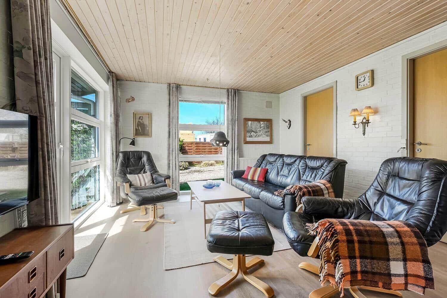 Wohnzimmer mit Ledersofa, zwei Sessel und großen Fenstern. Holzdecke und weiße Wände.