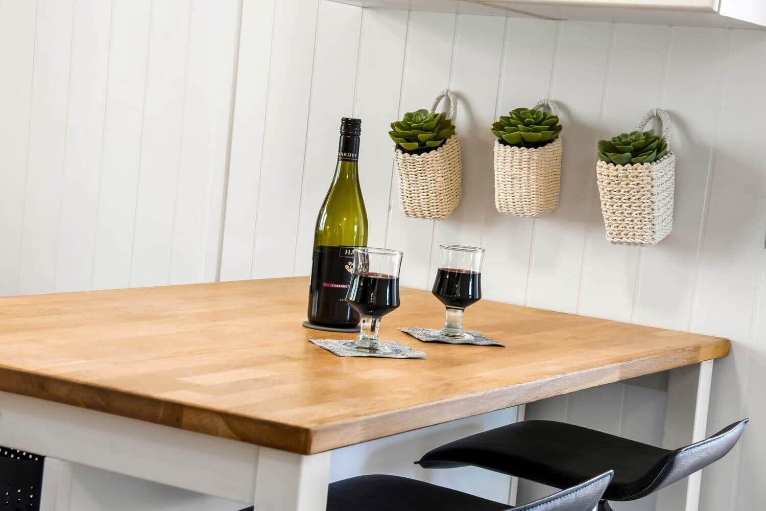 Holztisch mit Weinflasche und Gläsern, drei hängende Körbe mit Sukkulenten an weißer Wand.