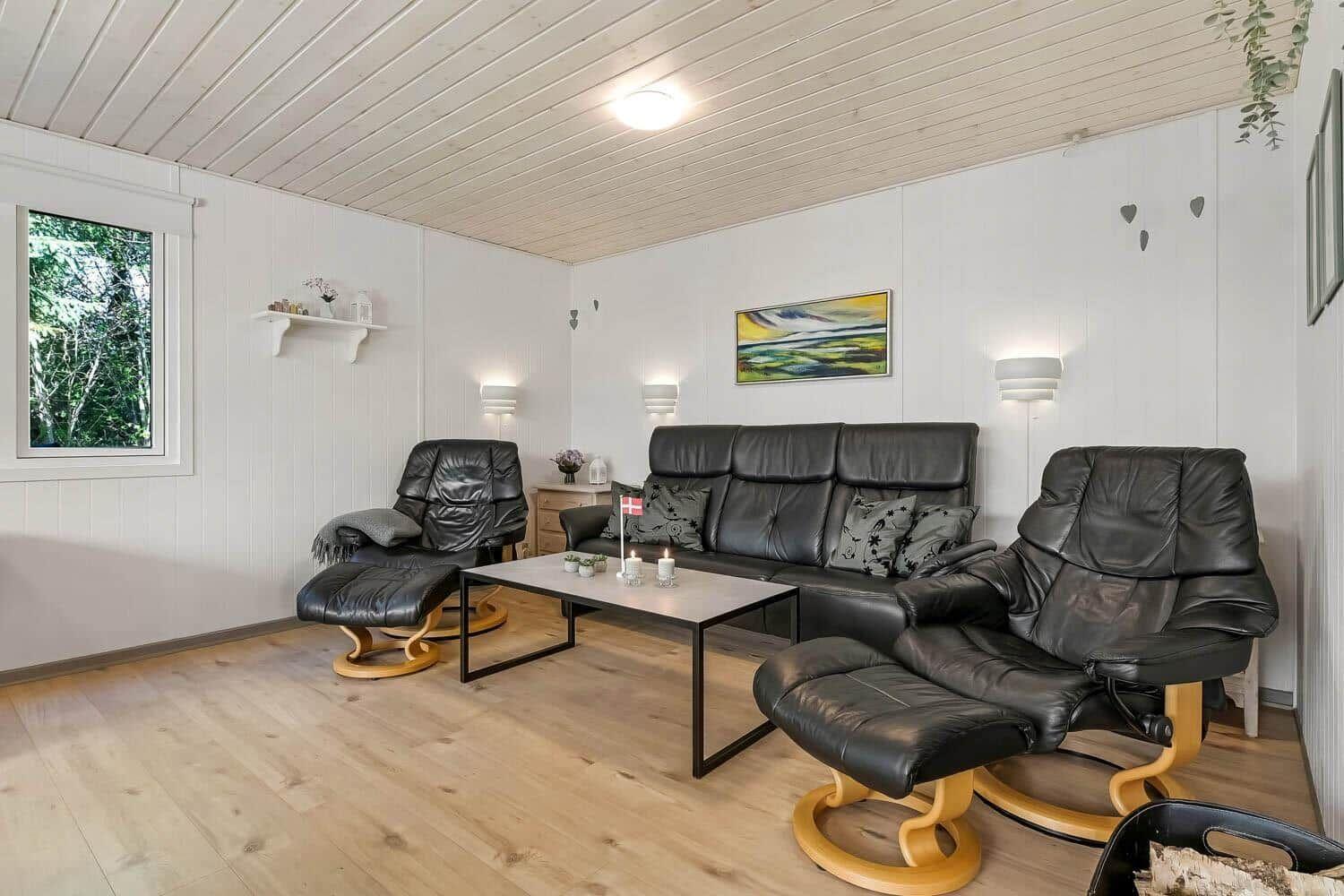 Wohnzimmer mit schwarzen Ledersofas, Holzboden und Fenster mit Blick auf Wald.