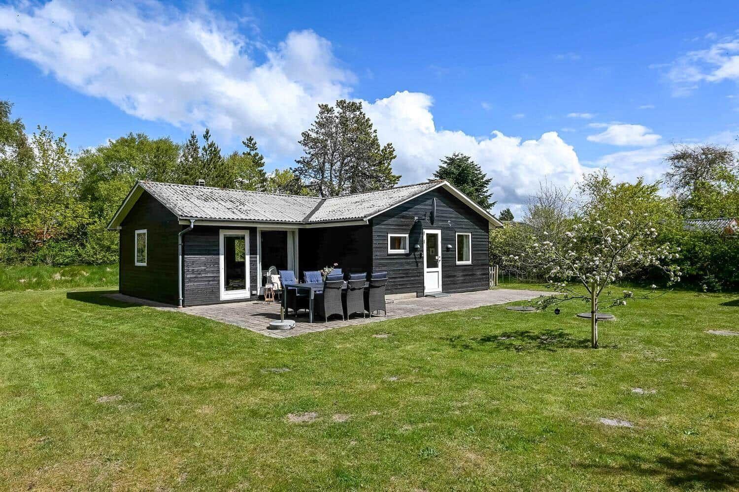 Ein dunkelgefärbtes Holzhaus mit Terrasse, Garten und Bäumen unter blauem Himmel.