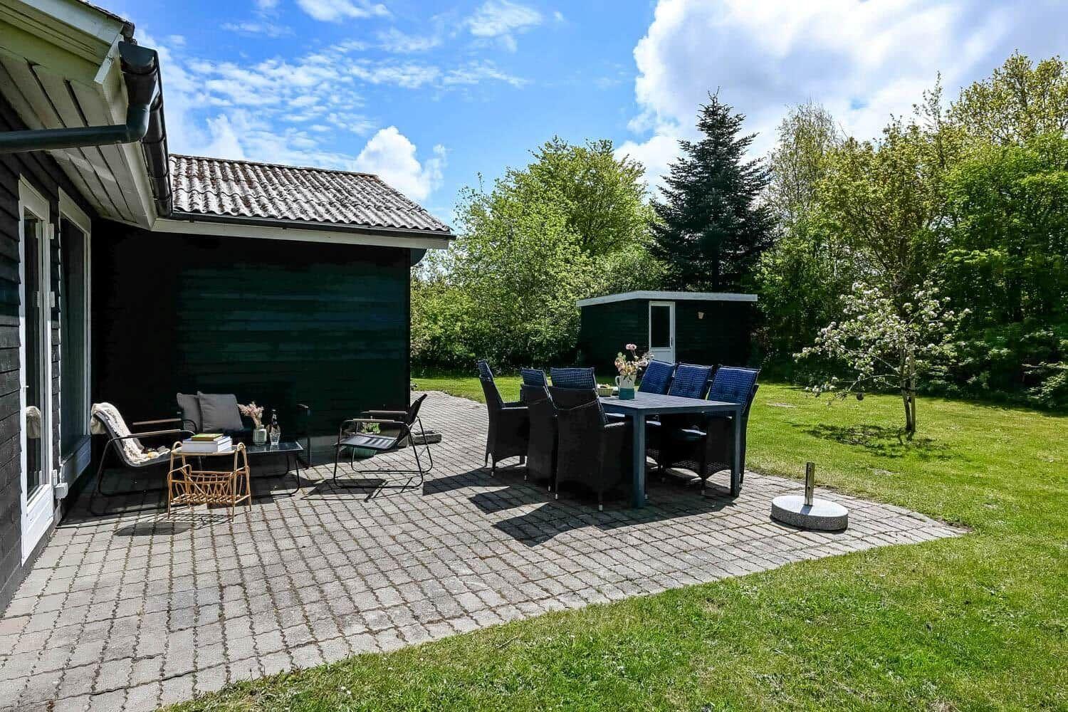 Terrasse mit Sitzgruppe und Esstisch im Garten. Hintergrund: grüne Bäume und ein kleines Gebäude.