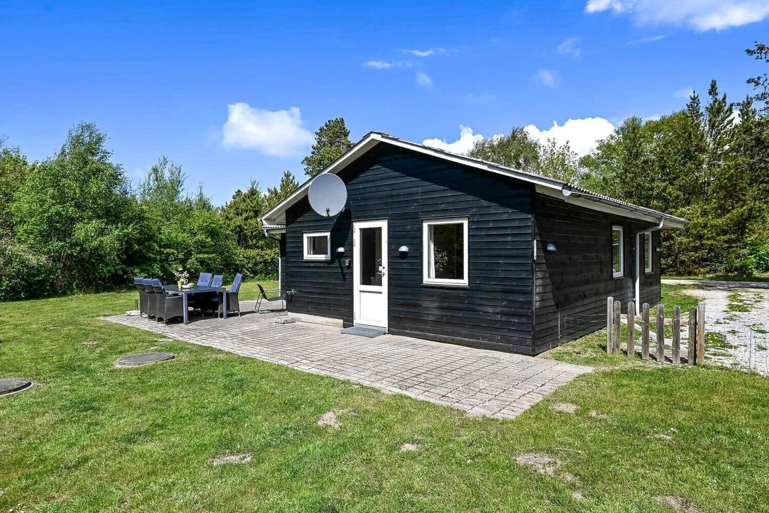 Schwarzes Holzhaus mit Terrasse, Garten und Satellitenschüssel. Umgeben von Bäumen und Gras.