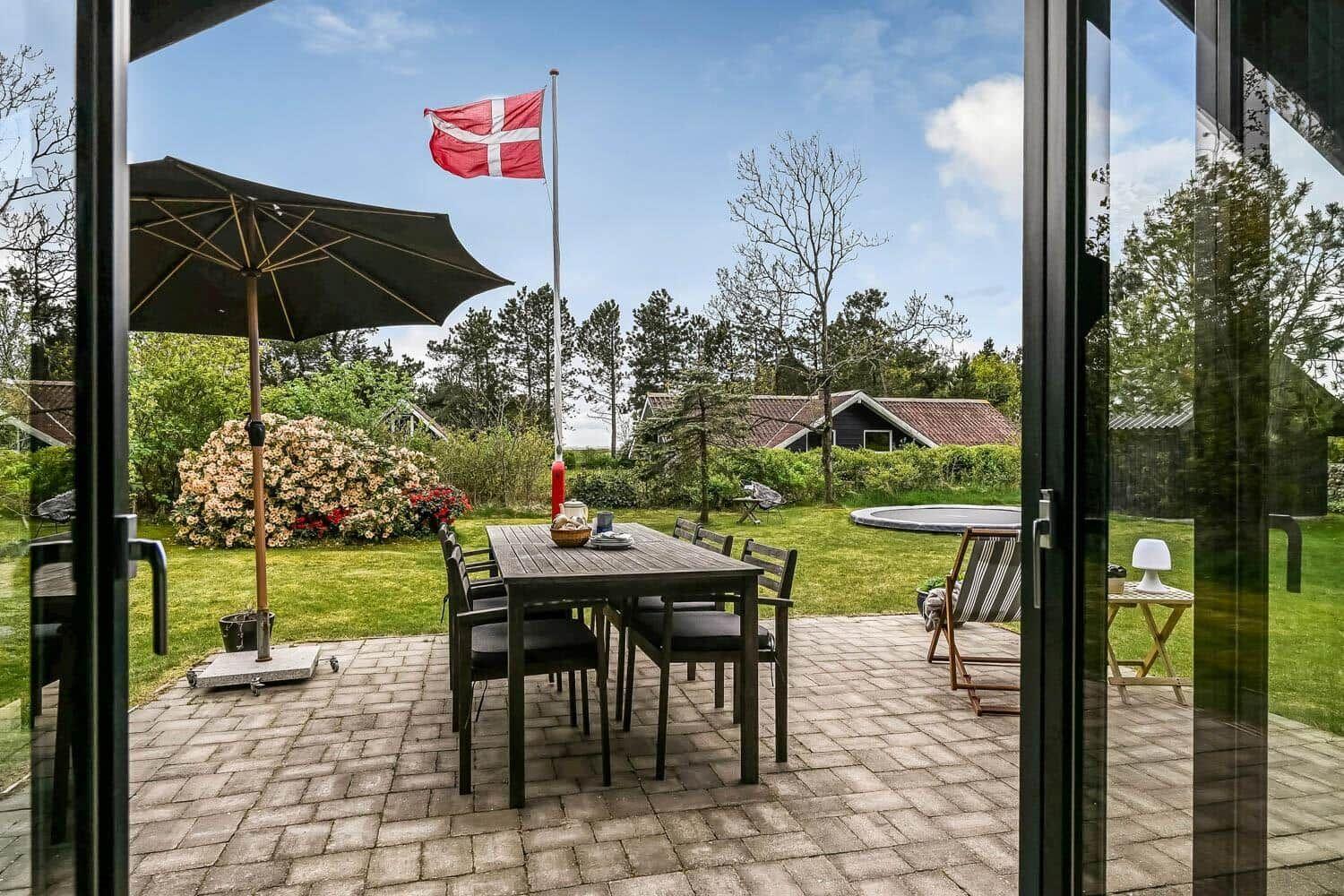 Terrasse mit Tisch, Stühlen und Sonnenschirm. Daneben Schwimmbad und dänische Flagge.