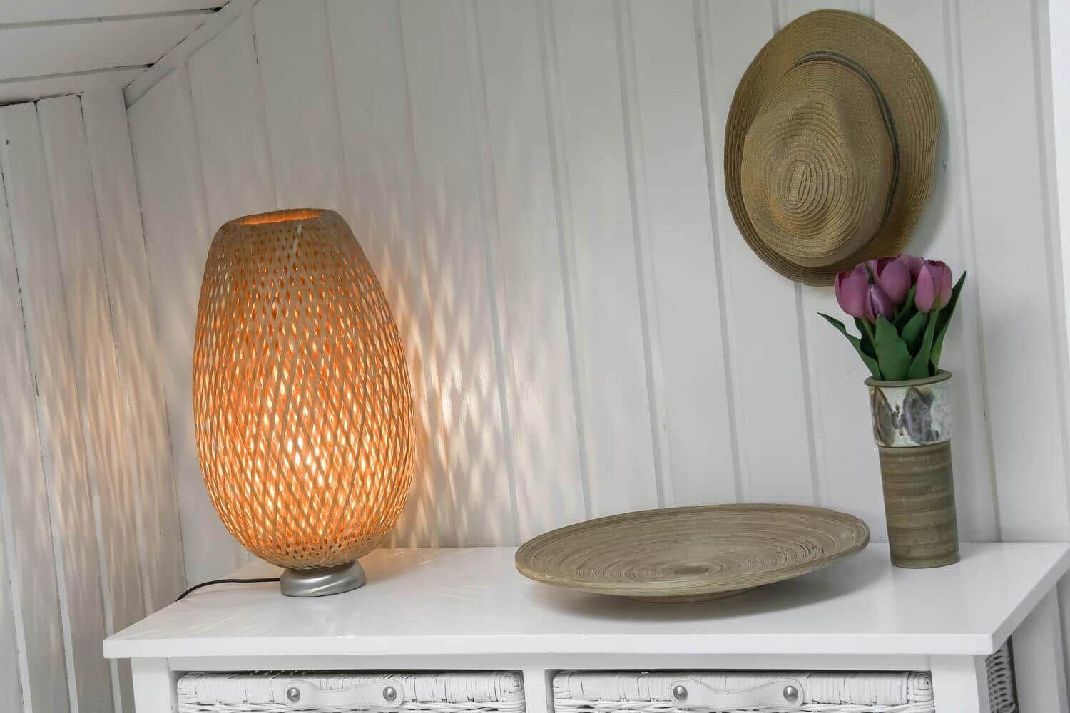 Auf einem weißen Tisch steht eine beleuchtete Lampe, eine Schale und eine Vase mit Tulpen. An der Wand hängt ein Hut.
