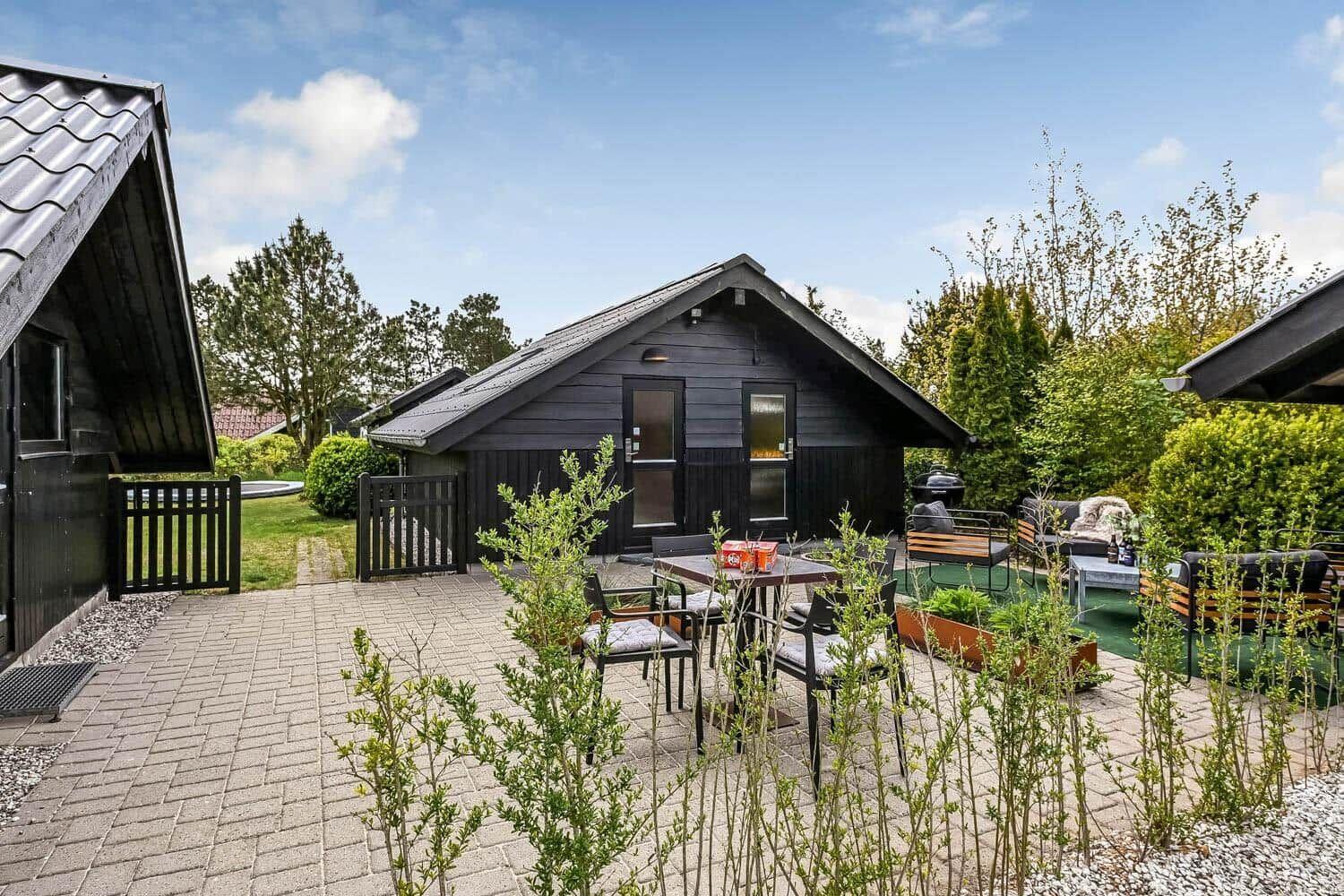 Schwarze Hütte mit Terrasse, Gartenmöbeln und Grillplatz. Umgeben von Bäumen und Grün.