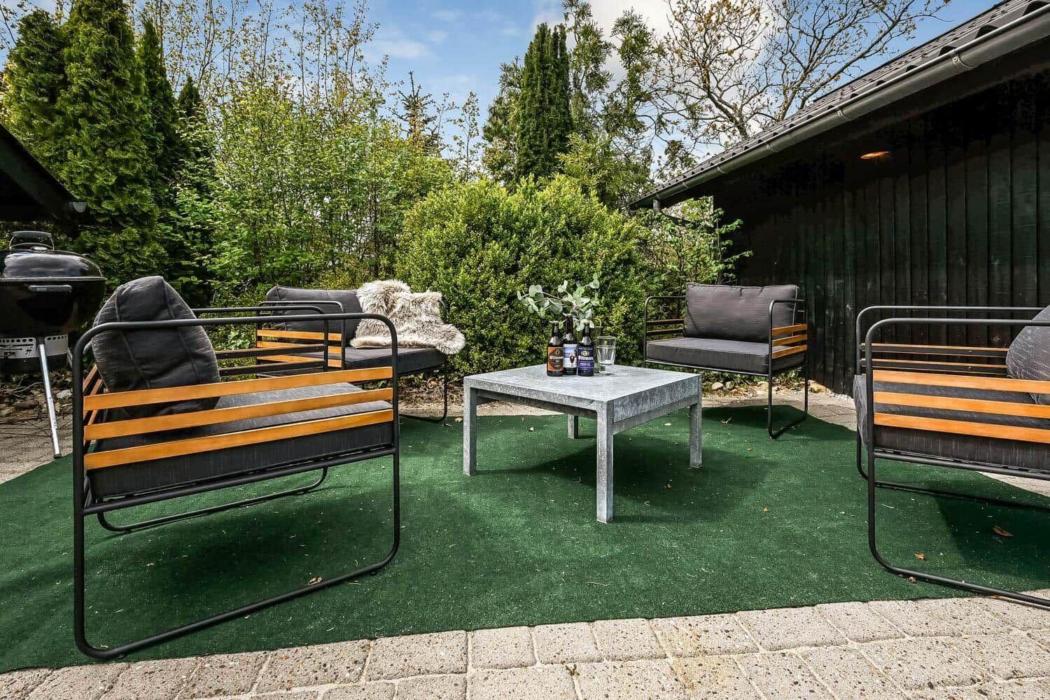 Terrasse mit Sitzgruppe, Tisch und Grill. Grünfläche und Bäume im Hintergrund.
