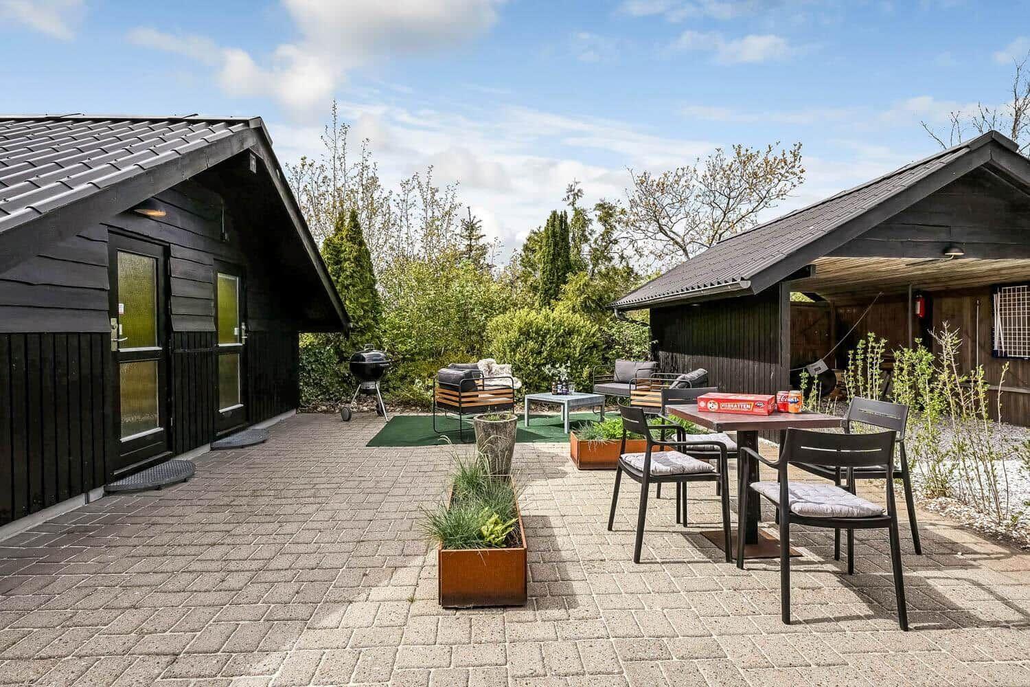 Terrasse mit Sitzgruppe, Grill und Tisch mit Stühlen. Zwei Häuser mit dunkler Fassade.