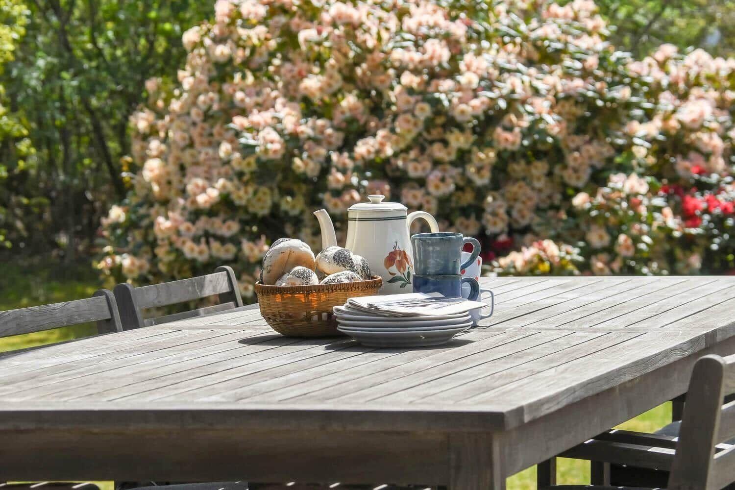 Holztisch mit Kanne, Tassen und Brot im Garten vor Blumenhecken.
