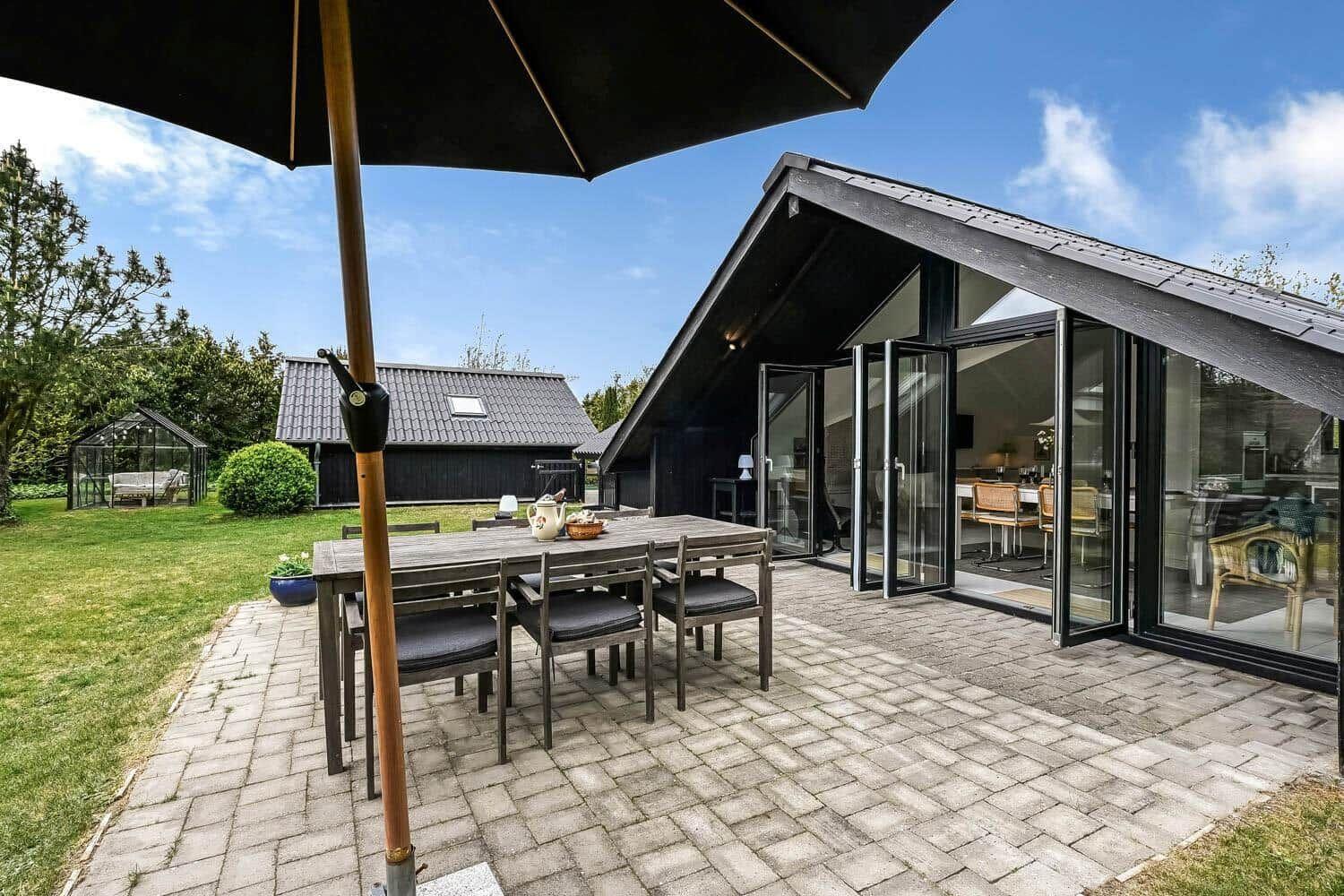 Terrasse mit Tisch, Stühlen und Sonnenschirm. Glasfront führt ins Haus. Grünfläche und Gartenhaus im Hintergrund.