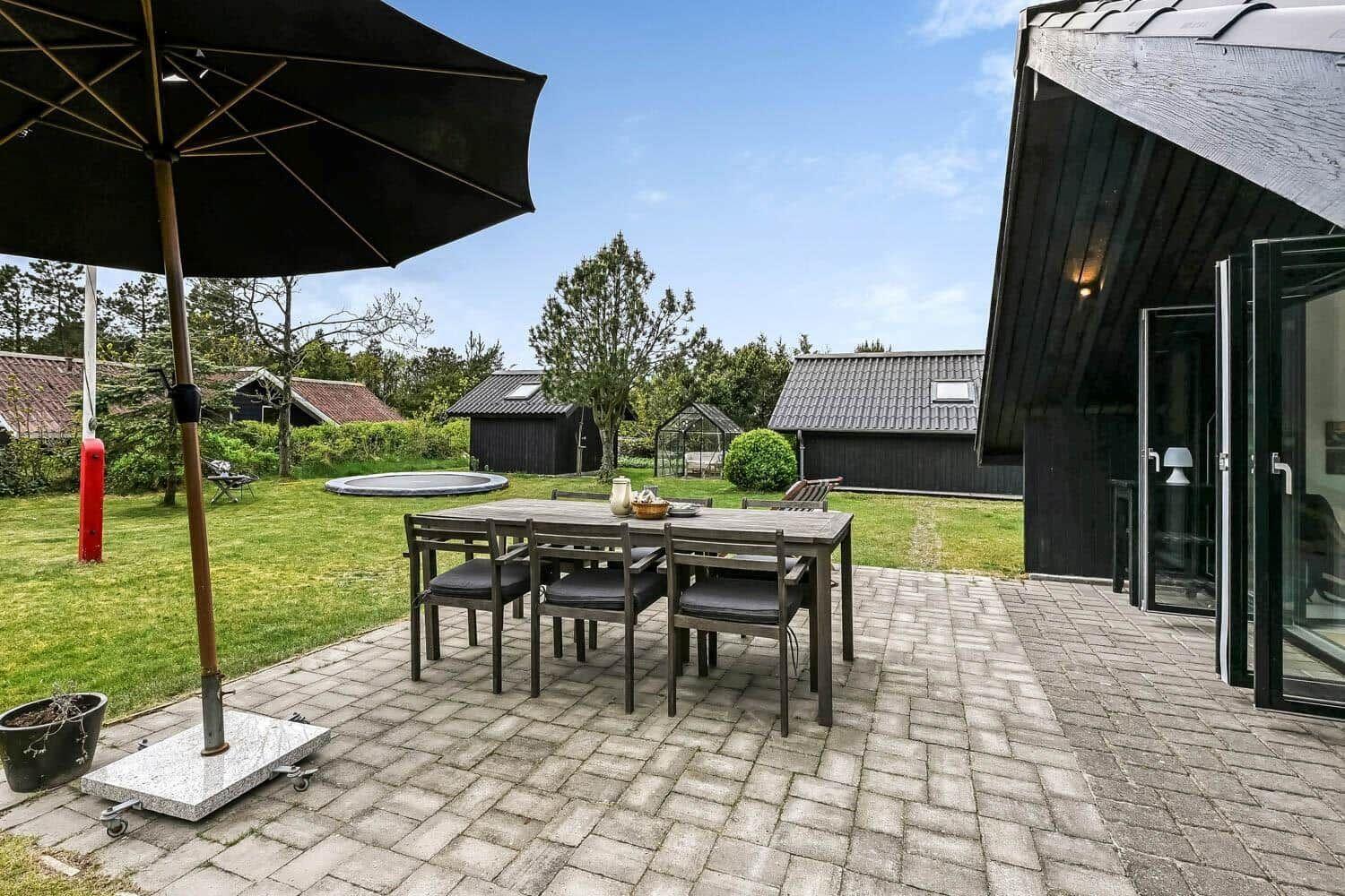 Terrasse mit Tisch, Stühlen und Sonnenschirm. Grünfläche und Pool im Hintergrund.