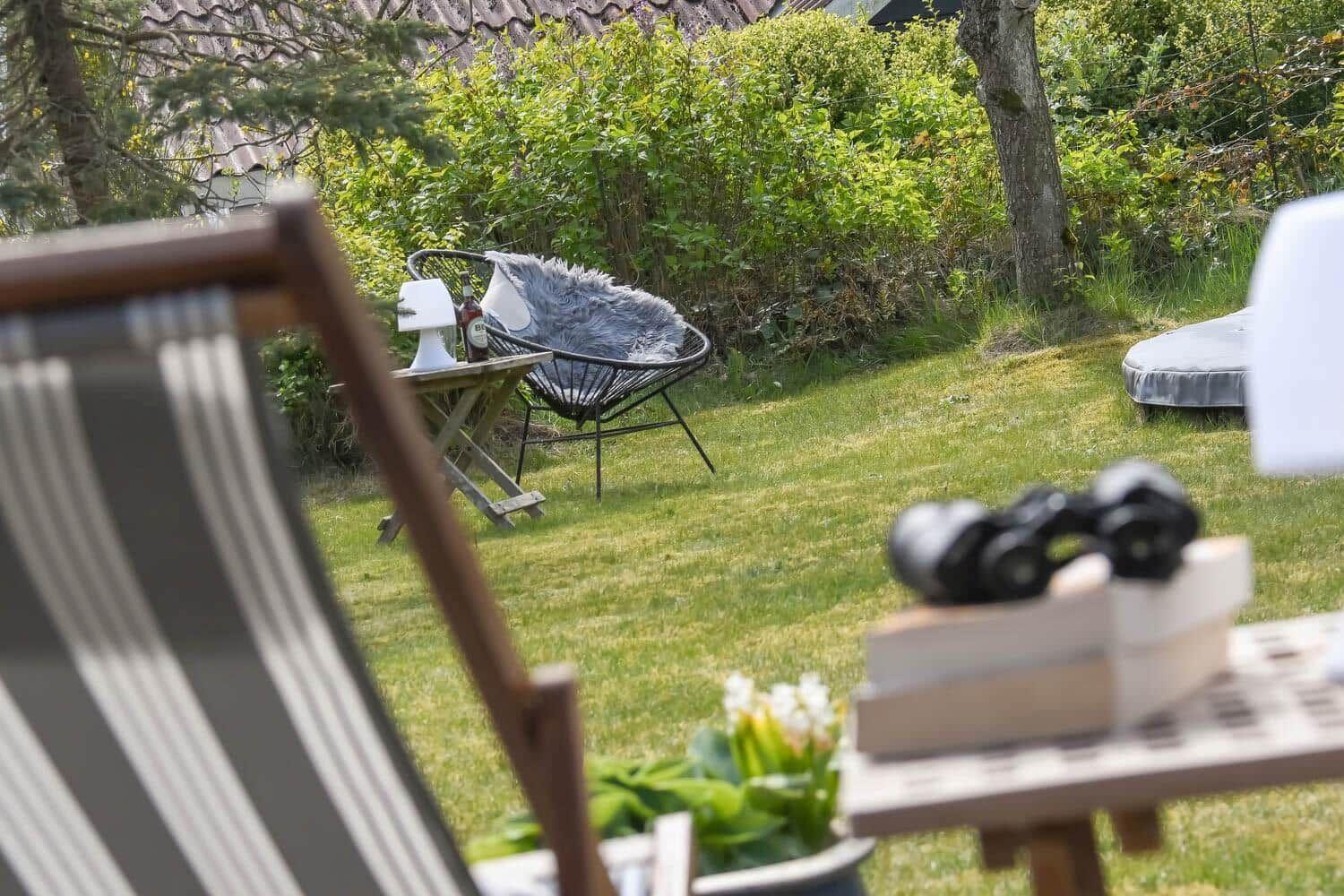Garten mit Liege, Tisch und Stühlen. Grünfläche, Bäume und Sträucher im Hintergrund.
