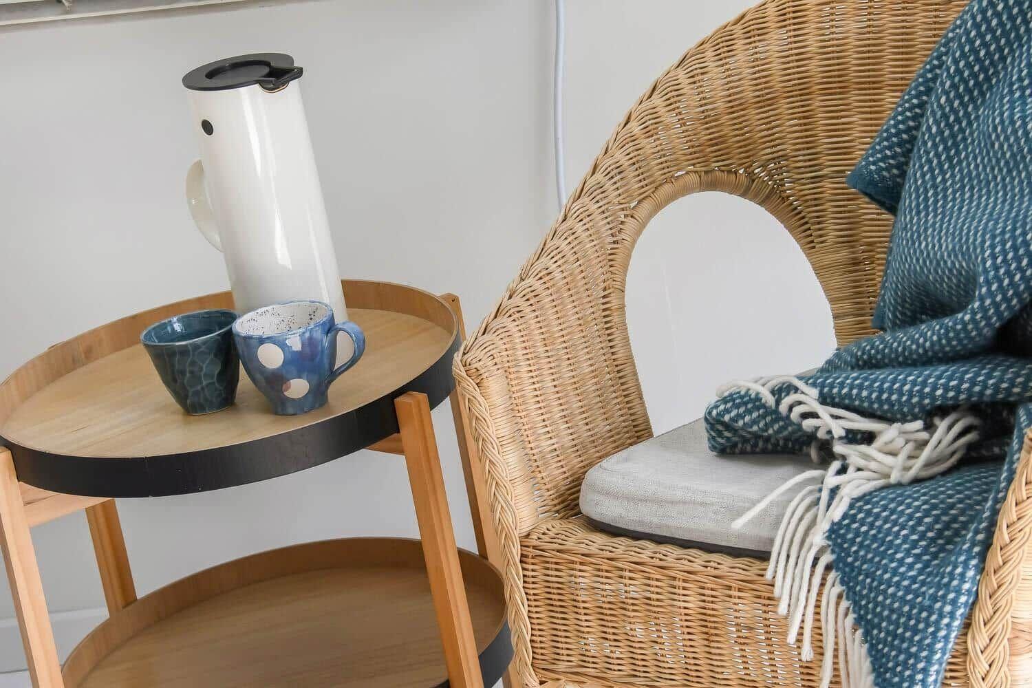 Rattan-Stuhl mit Kissen und Decke neben einem Holztisch mit Thermoflasche und Tassen.