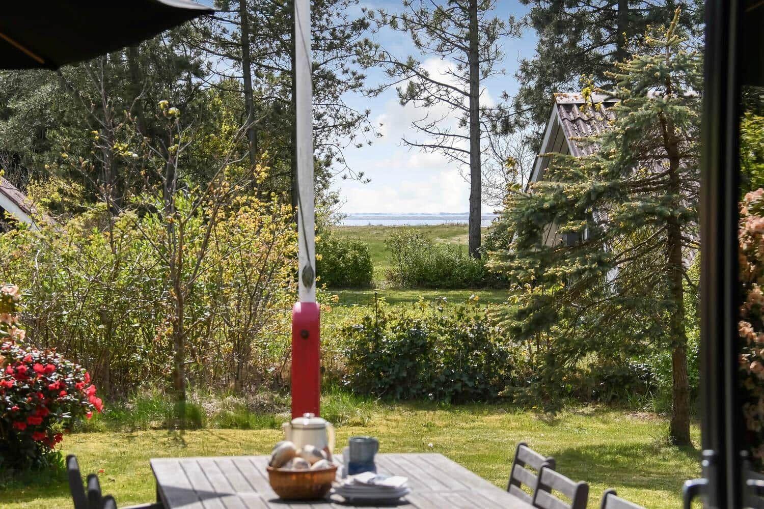 Terrasse mit Tisch und Stühlen, Blick auf Grünflächen und Wasser durch Bäume.