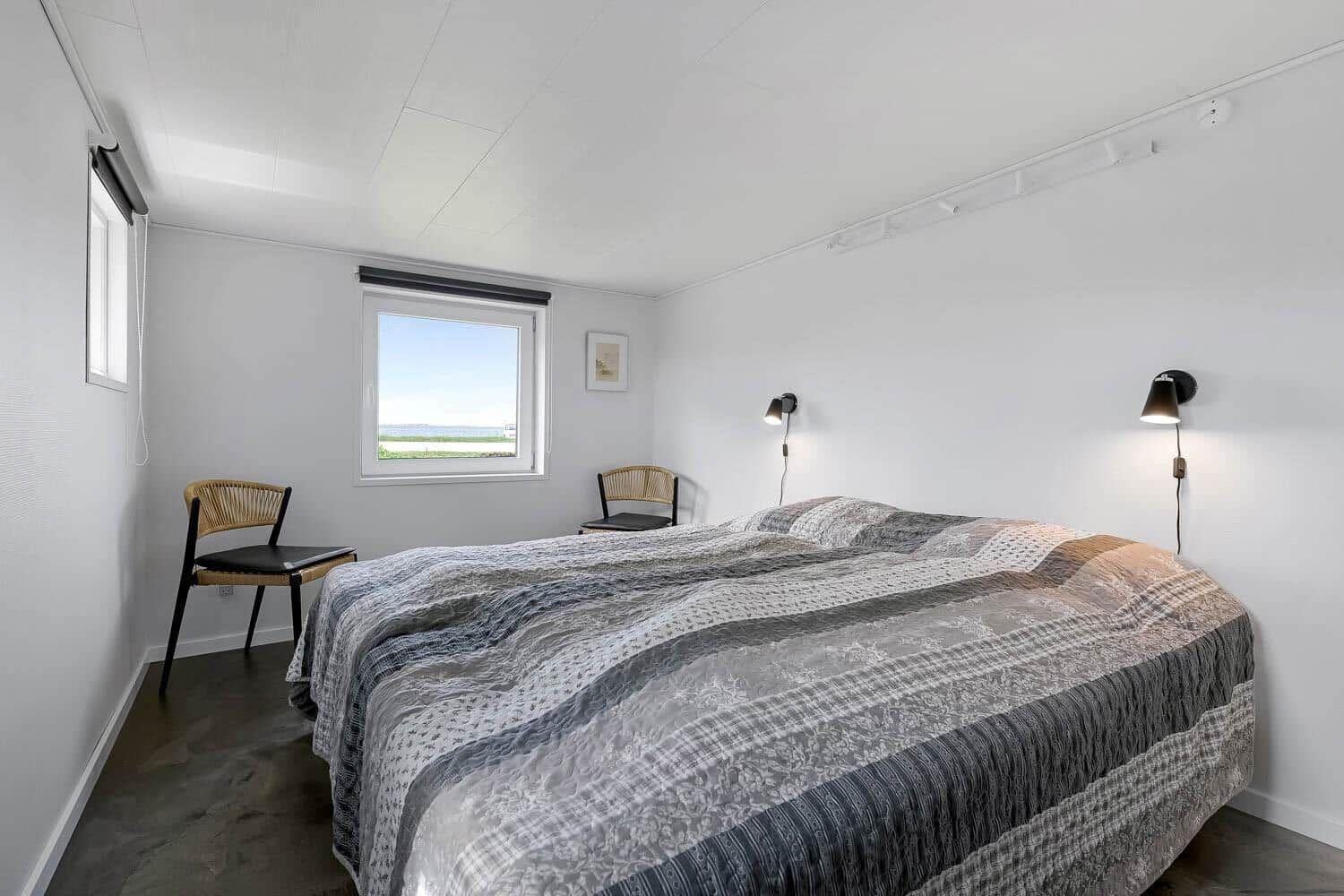 Schlafzimmer mit Bett, zwei Stühlen und Fensterblick auf Wasser.