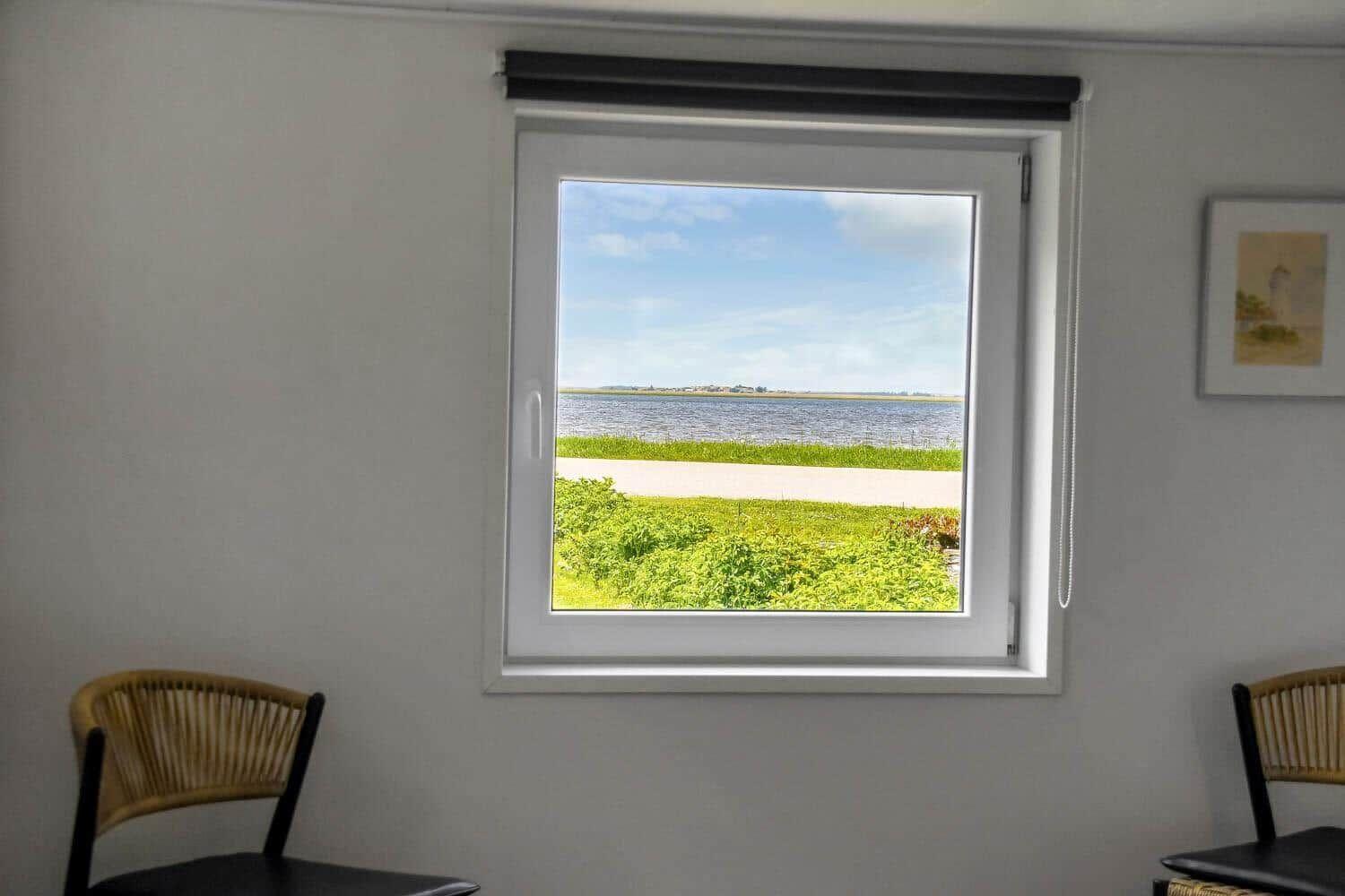 Durch das Fenster sieht man einen See mit Grünflächen und einem Weg. Zwei Stühle stehen im Raum.