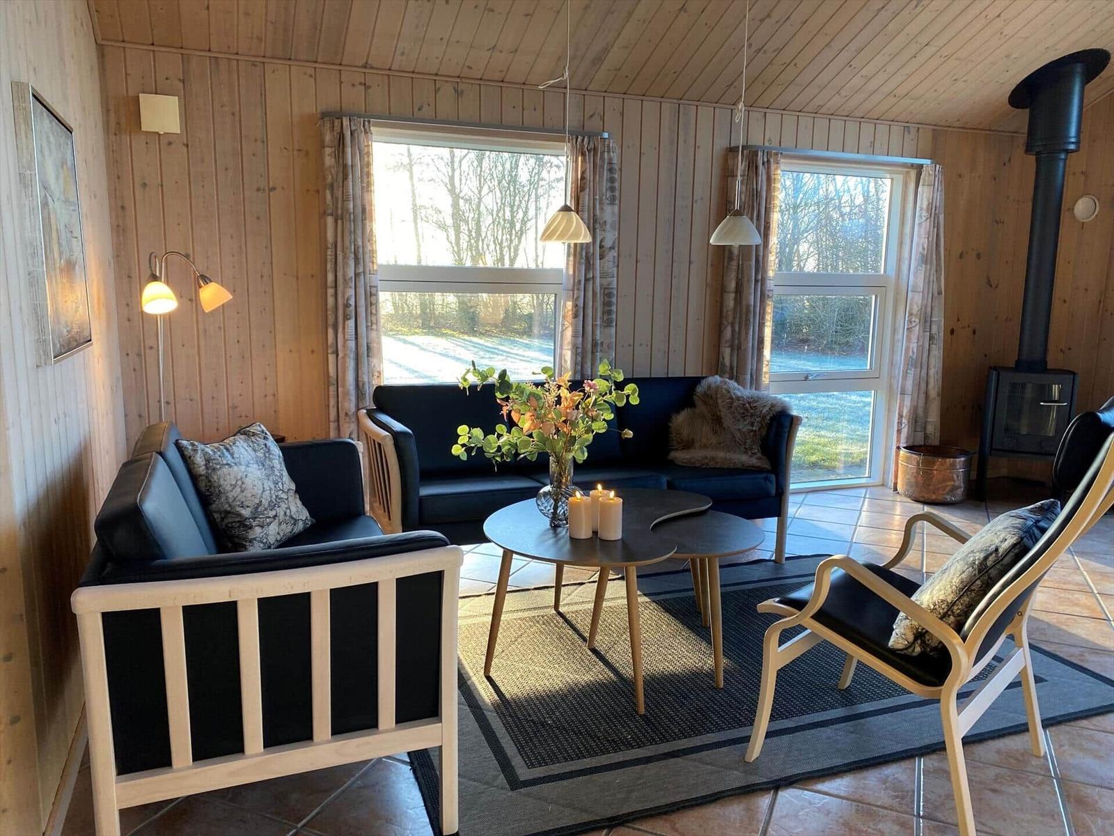 Wohnzimmer mit Holzwand, Sofa, Stühlen und Kaminofen. Fenster mit Blick auf Wald.