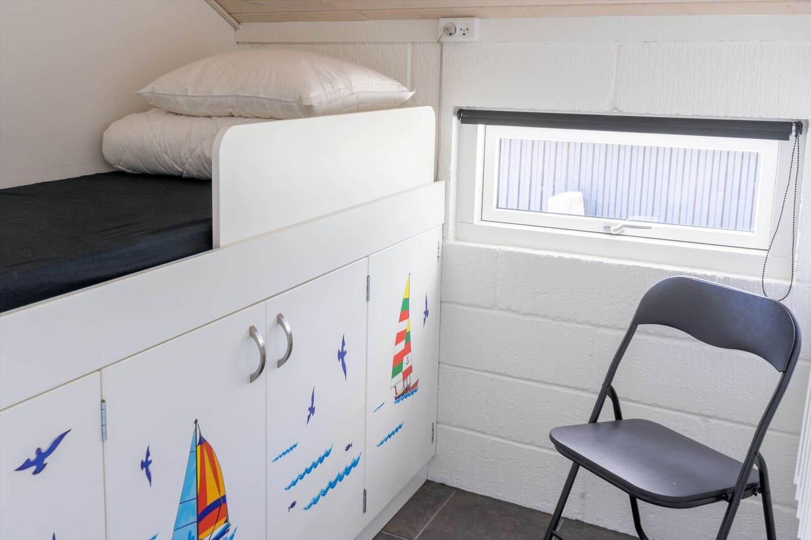 Obere Schlafcouch mit Matratze und Kissen. Unterer Teil ist Schrank mit Segelboot-Aufklebern. Fenster und Stuhl daneben.