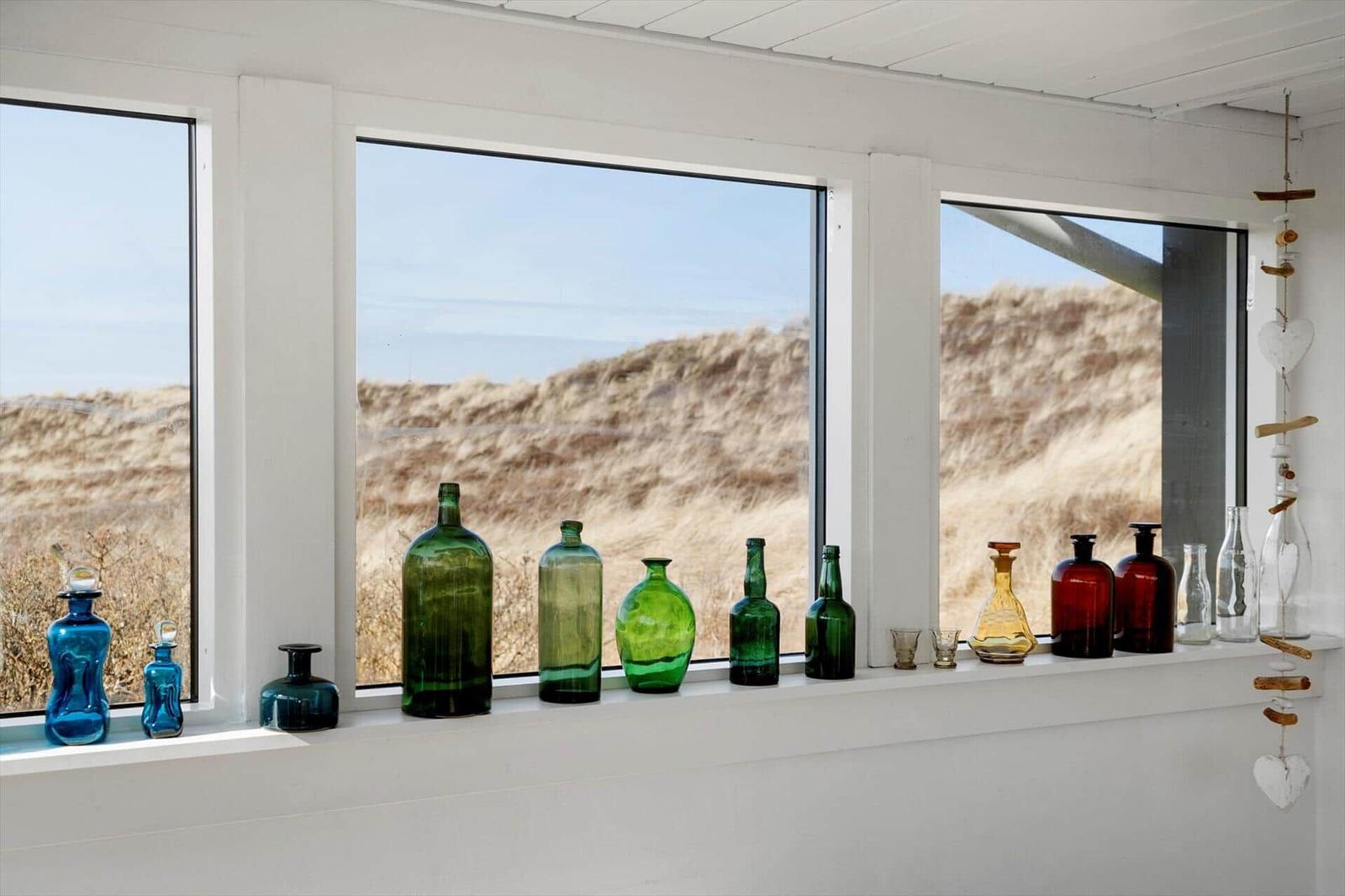 Fensterbank mit bunten Glasflaschen und Blick auf Dünen.