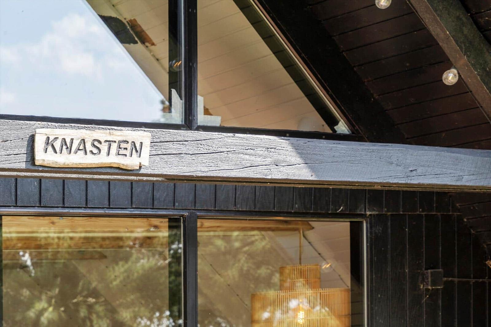 Haus mit Schild KNASTEN, dunkle Holzwand und Fenster mit Blick ins Innere.