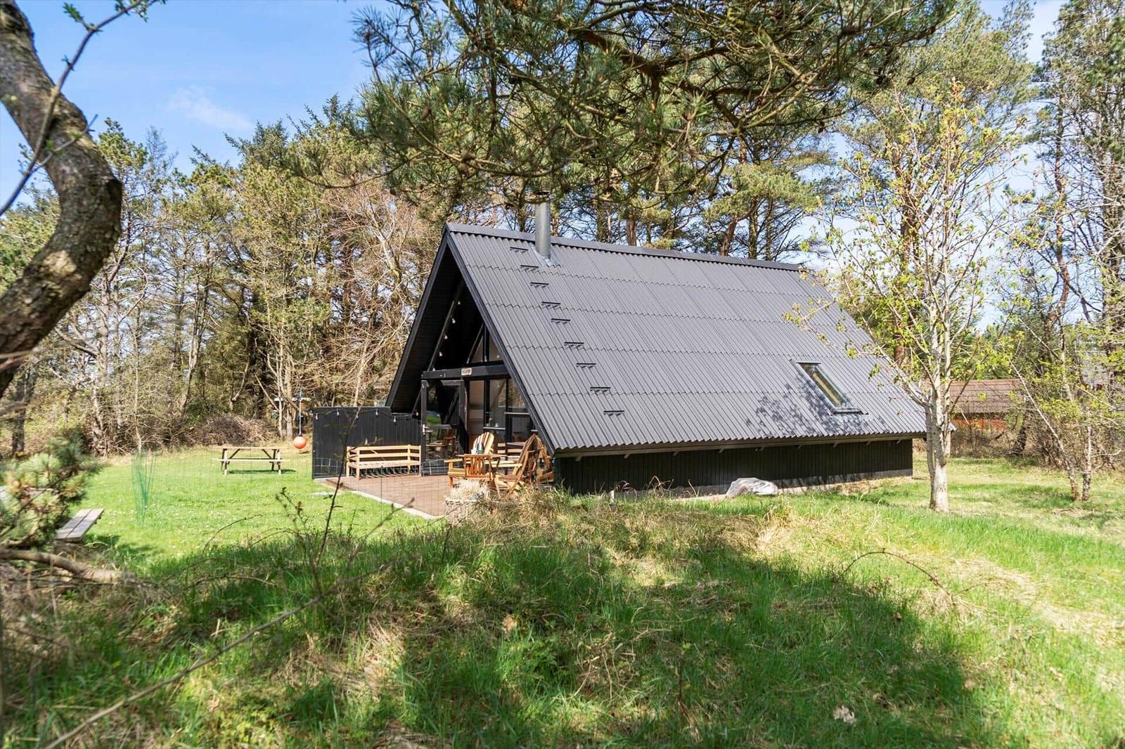 Moderne Holzhütte mit dunklem Dach und Terrasse im Wald. Grasfläche und Bäume umgeben das Haus.