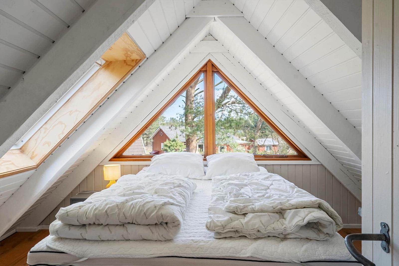 Schlafzimmer mit Dachschrägen, großem Fenster und Bett mit weißen Decken.