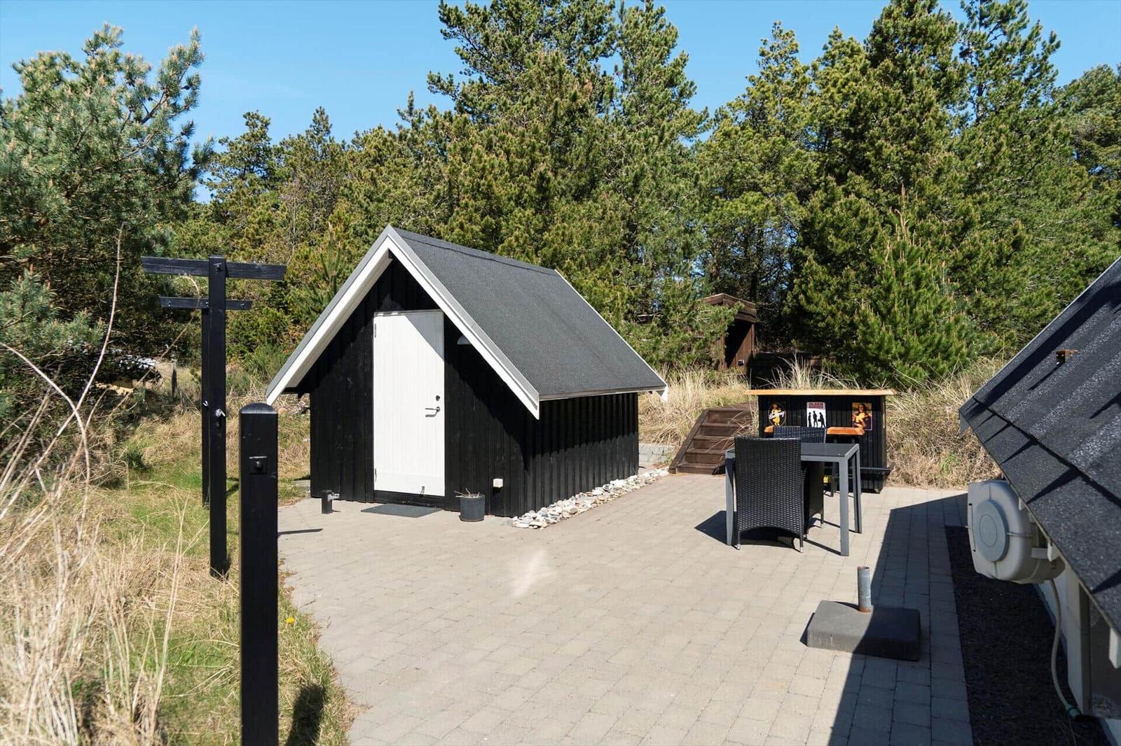 Schwarze Hütte mit weißer Tür, Terrasse und Gartenmöbeln zwischen Bäumen.