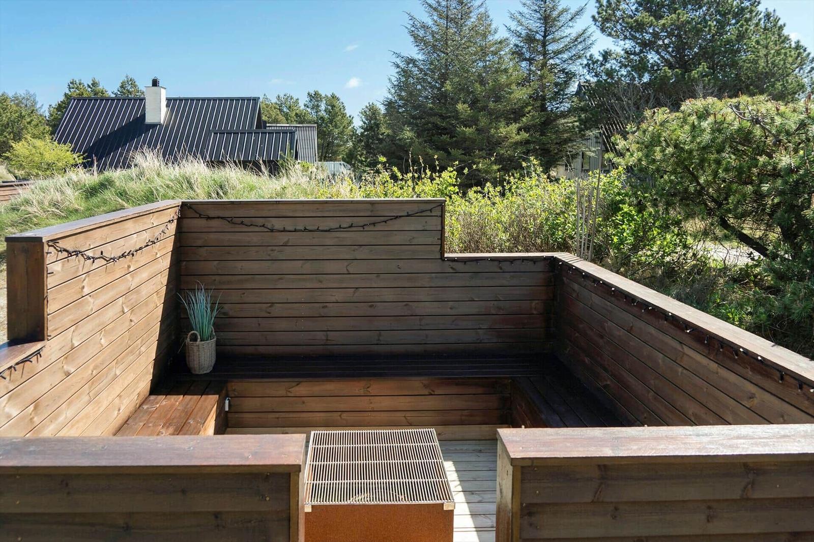 Holzterrasse mit Sitzgelegenheit und Grill. Im Hintergrund sind Bäume und ein Haus zu sehen.