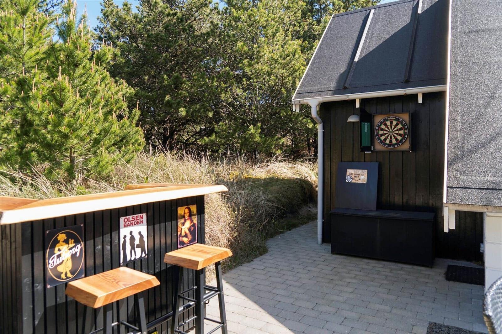Außenbereich mit Dartscheibe, Bar und Holzstühlen. Hintergrund mit Nadelbäumen und Gras.