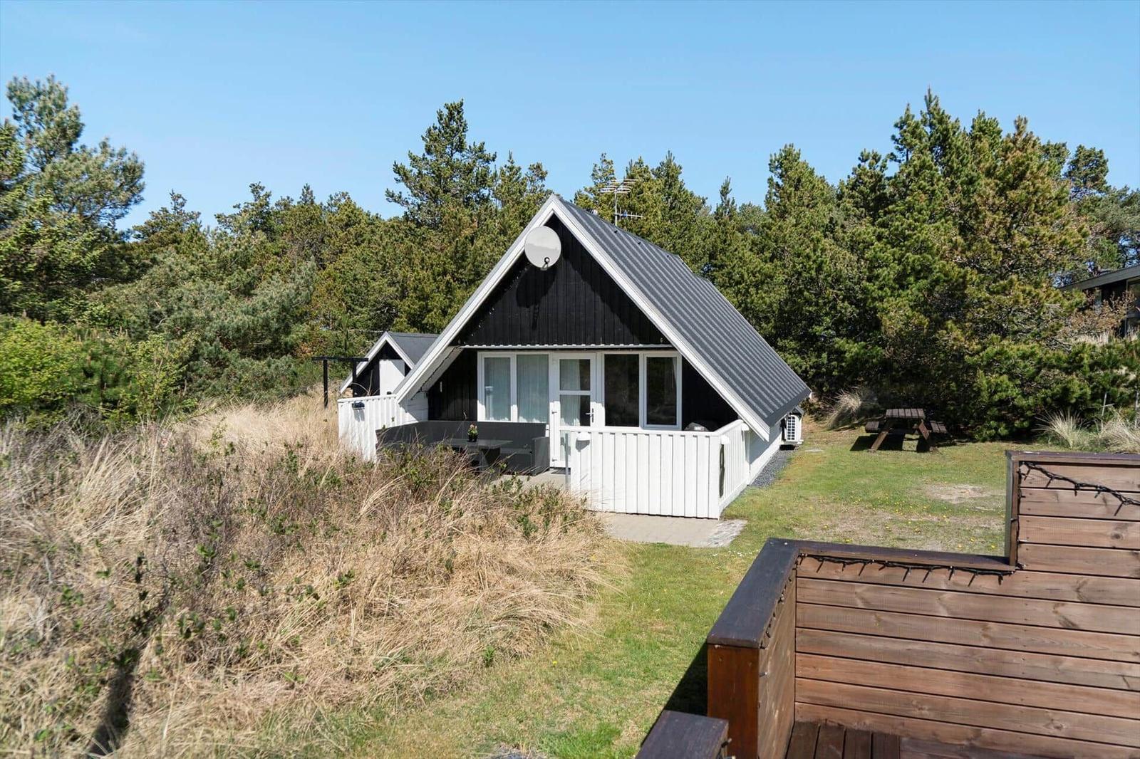Ein A-Fachhaus mit weißem Holzgeländer und dunklem Dach. Umgeben von Gras und Bäumen.