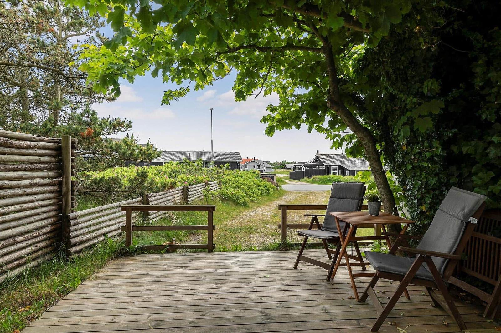 Holzterrasse mit zwei Stühlen und Tisch unter Bäumen. Blick auf Grünflächen und Häuser.